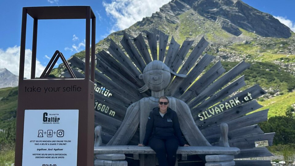 A person is sitting on a large wooden figure wearing sunglasses. In the background, there are mountains and a clear blue sky.