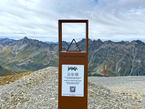 An information sign on a mountain path with a view of the surrounding mountains. The sky is cloudy and the landscape is rocky.