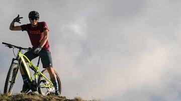 A cyclist poses with his mountain bike on a hill. In the background, clouds can be seen in the sky.