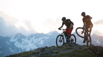 Two mountain bikers are standing on a hill, enjoying the view of the snow-covered mountains. The sky is cloudy, and the scene has an adventurous feel.