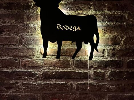 A stylized silhouette of a cow on a brick wall. Below it, "Bodega" is written in curvy script.