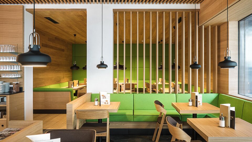 Ein modernes Café mit Holzmöbeln und einem klaren, minimalistischen Design. Die Wände sind mit einem lebendigen Grün akzentuiert, und es gibt stilvolle Pendelleuchten.