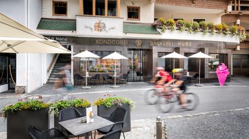 Eine belebte Straße mit einem Konditorei-Café und Außenbereich. Radfahrer fahren vorbei, während Tische und Sonnenschirme zum Verweilen einladen.