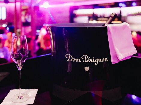 A bottle of Dom Pérignon in an elegant ice bucket is ready. In the background, a festive atmosphere is visible with a blurred audience.
