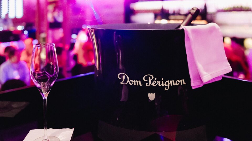 A bottle of Dom Pérignon in an elegant ice bucket is ready. In the background, a festive atmosphere is visible with a blurred audience.