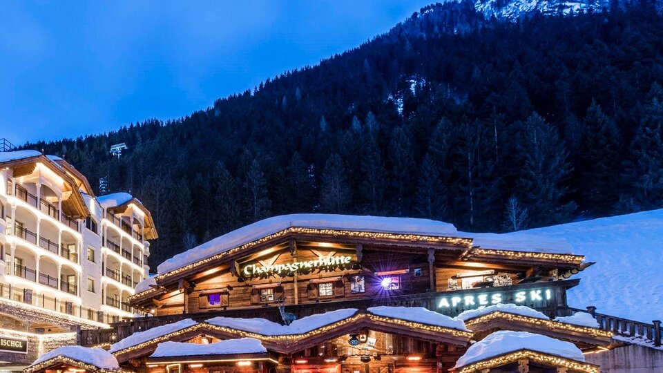 Eine gemütliche Berghütte im Schnee mit Lichtern, die in der Dämmerung leuchten. Im Hintergrund sind schneebedeckte Berge und Wälder zu sehen.