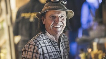 Ein lächelnder Mann mit einem Hut und einer karierten Bluse. Im Hintergrund sind verschwommene Lichtquellen zu sehen.