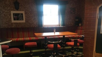 A cozy bar area with red stools and a table. In the background, there is a decorative wall and a window with daylight.