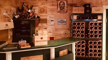A cozy wine cellar with wooden walls displaying wine boxes. In the background, there is a shelf with wine bottles and a refrigerator.