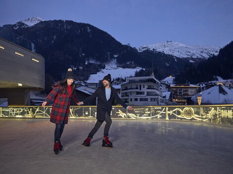 A couple is enjoying a lively round on an ice rink in the mountains. In the background, snow-covered peaks and cozy houses can be seen.