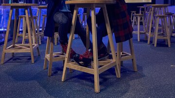 Two people are sitting at high tables in a modern room with blue lighting. They are wearing warm clothing and enjoying drinks.