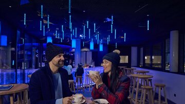 A café with two people sitting in warm winter clothing and chatting. The environment is illuminated in blue light, and they are enjoying hot drinks.