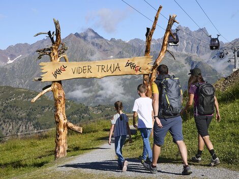 A hiking group is walking along a picturesque path to a sign that reads "Vider Truja". In the background, majestic mountains and a cable car can be seen.