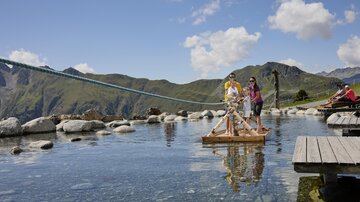 A tranquil mountain lake with people standing on a raft. In the background, green mountains and a blue sky with some clouds are visible.