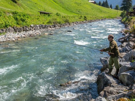 A man is fishing at a clear, flowing river. Surrounded by green meadows and trees, the landscape offers a serene natural backdrop.