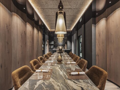 An elegant dining area with a long table and cozy chairs. The atmosphere is modern and stylish, illuminated by large lamps.