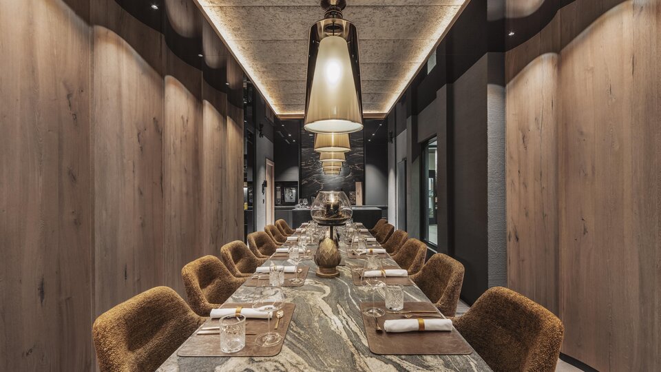 Ein eleganter Restaurantbereich mit einem langen Tisch und gemütlichen Stühlen. Die Atmosphäre ist modern und stilvoll, beleuchtet von großen Lampen.
