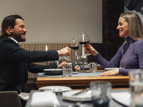 An elegant dinner couple toasts with red wine. The table is set and the atmosphere feels cozy.