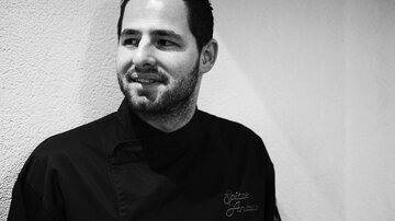 A smiling chef stands by a wall. He is wearing a dark chef's jacket and looks friendly and relaxed.