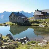 Eine malerische alpine Landschaft mit zwei Hütten am Ufer eines ruhigen Sees. Im Hintergrund erheben sich majestätische Berge unter einem klaren Himmel.