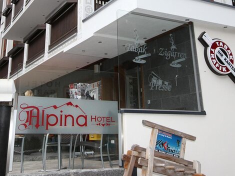 Ein modernes Hotel mit der Aufschrift "Alpina Hotel" ist zu sehen. Die Fassade hat große Fenster und zeigt einen einladenden Eingangsbereich.