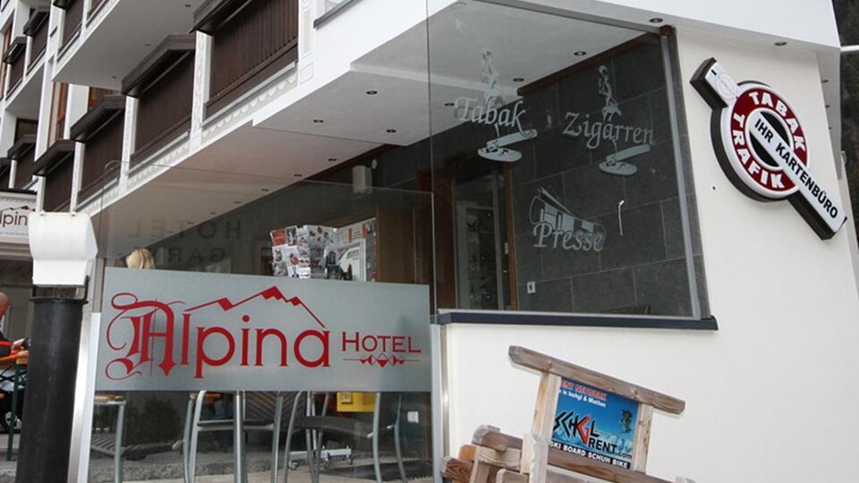 Ein modernes Hotel mit der Aufschrift "Alpina Hotel" ist zu sehen. Die Fassade hat große Fenster und zeigt einen einladenden Eingangsbereich.