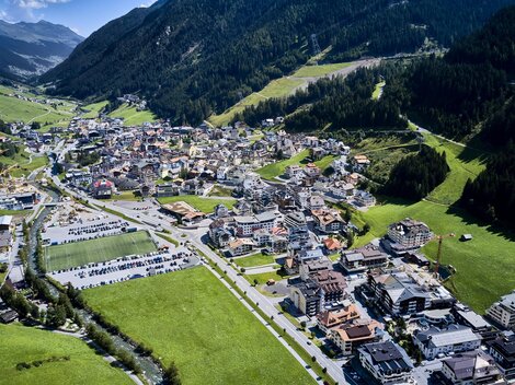 A picturesque alpine town in a green valley, surrounded by mountains. The buildings are closely packed, and there are many parking spaces and streets.