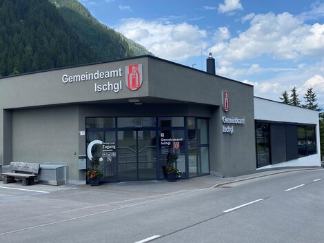 The image shows the municipality office of Ischgl, a modern building with large windows. In the background, mountains and a blue sky can be seen.