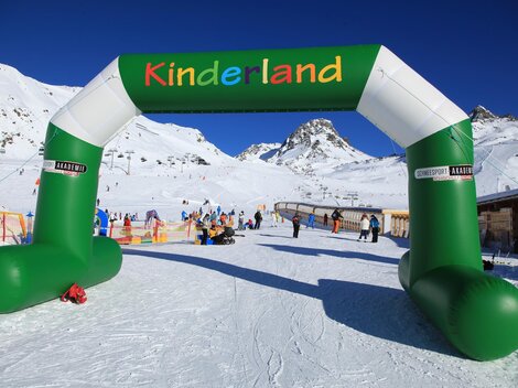 A colorful arc with the inscription "Kinderland" stands in a snowy ski resort. In the background, ski lifts and skiers can be seen.