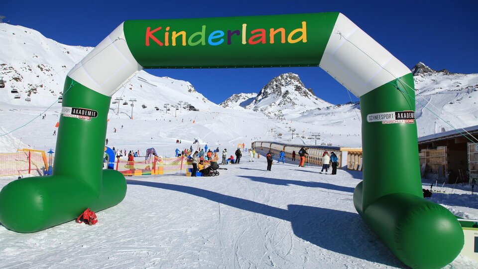 Ein bunter Bogen mit dem Schriftzug "Kinderland" steht in einem verschneiten Skigebiet. Im Hintergrund sind Schlepplifte und Skifahrer zu sehen.