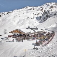 Ein belebtes Skigebiet mit einem großen Restaurant inmitten schneebedeckter Berge. Viele Menschen genießen die Sonne und die Winteraktivitäten.