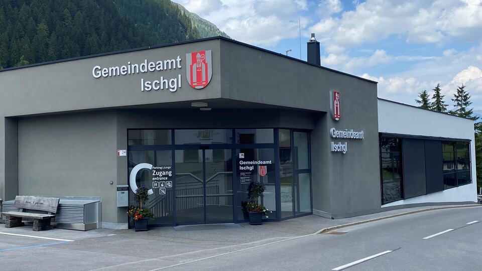 Ein modernes Gebäude des Gemeindeamts Ischgl mit klarer Beschilderung. Im Hintergrund sind grüne Berge und ein blauer Himmel zu sehen.