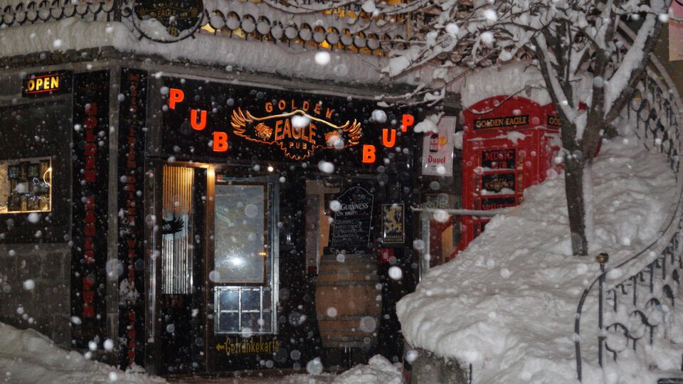 A cozy pub named "Golden Eagle" is hidden in the snow. The snowflakes fall thickly as the warm light of the entrance seems inviting.