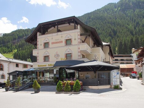 A picturesque hotel in a mountainous environment with green mountains in the background. In front of the hotel, there is a paved area with tables and chairs.