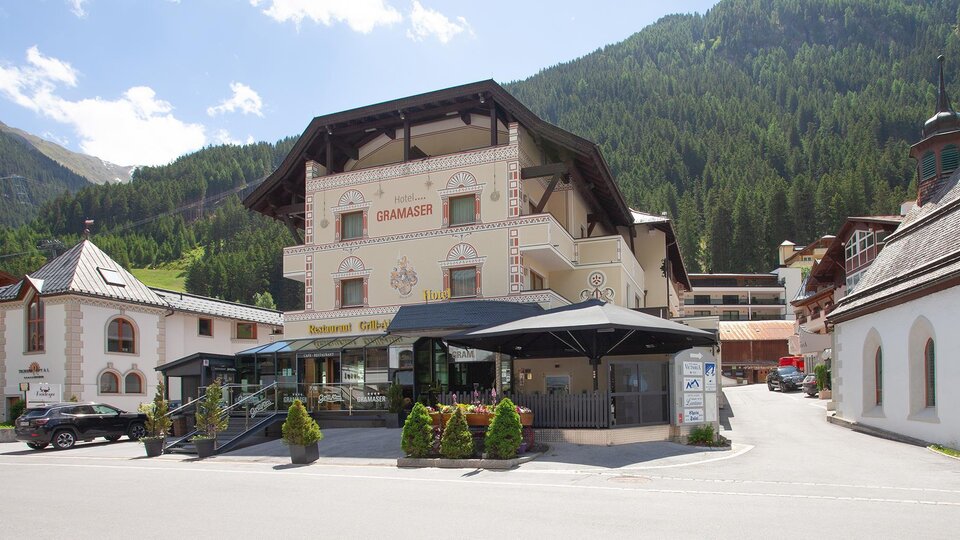 A picturesque hotel in a mountainous environment with green mountains in the background. In front of the hotel, there is a paved area with tables and chairs.