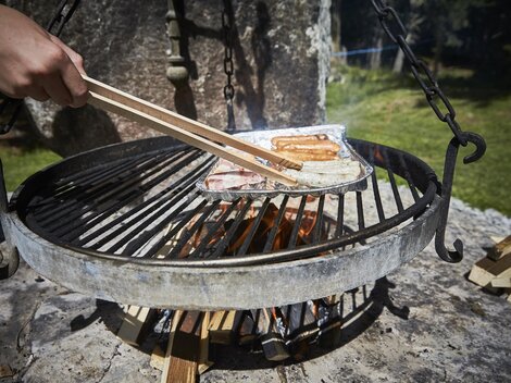 A grill with sizzling sausages is being prepared over an open flame. In the background, green grass and nature can be seen.