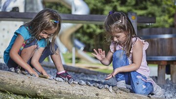 Two children are playing outside and collecting stones on a tree trunk. In the background, a playground and green meadows can be seen.