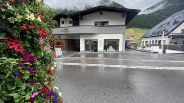 A modern building in a rural setting, surrounded by mountains. Colorful flowers hang on the left side, and it seems like it might rain.