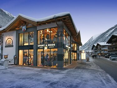 Ein modernes Geschäft in den Alpen, umgeben von schneebedeckten Bergen. Die Fassade ist großflächig verglast und einladend.