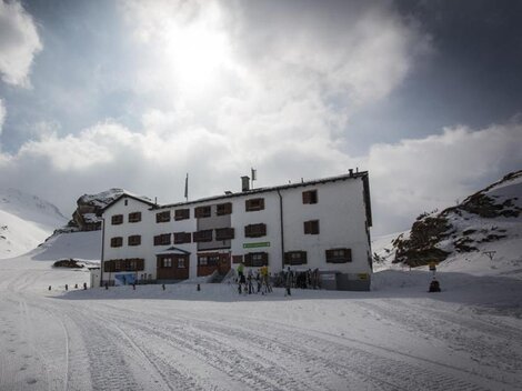 A white building in the snow, surrounded by mountains and clouds. People can be seen near the house, which radiates a cozy atmosphere.