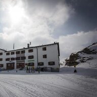 Ein weißes Gebäude im Schnee, umgeben von Bergen und Wolken. Menschen sind in der Nähe des Hauses zu sehen, das eine gemütliche Atmosphäre ausstrahlt.