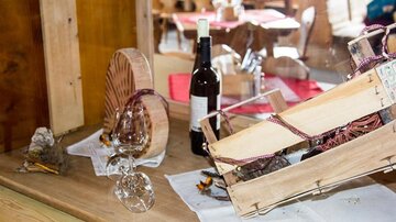 A decorative table with an open wine basket, a bottle of wine, and two wine glasses. In the background, simple wooden furniture and a cozy atmosphere are visible.