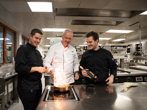 Three chefs are standing in a modern kitchen, preparing a flambéed dish. They appear focused and enthusiastic about the art of cooking.