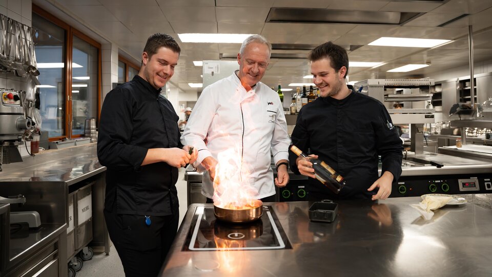 Three chefs are standing in a modern kitchen, preparing a flambéed dish. They appear focused and enthusiastic about the art of cooking.