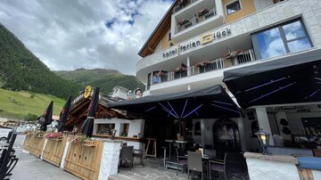 A cozy hotel with an outdoor area and terrace furniture. In the background, green mountains and a cloudy sky are visible.