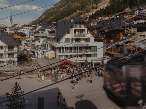 A bustling place in the mountains with a hotel and many people. In the background, wooden buildings and a cable car can be seen.