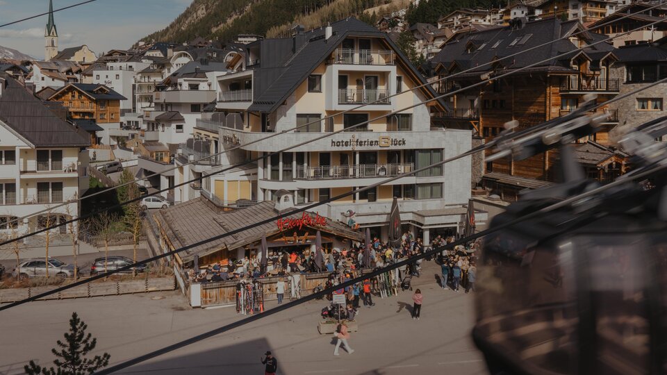 A bustling place in the mountains with a hotel and many people. In the background, wooden buildings and a cable car can be seen.
