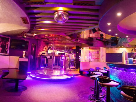 A stylish club with a lit bar area and a dance floor in the middle. The colorful lights create a vibrant atmosphere.