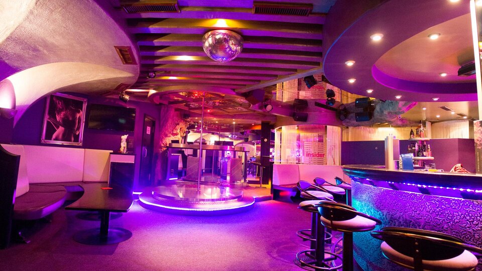 A stylish club with a lit bar area and a dance floor in the middle. The colorful lights create a vibrant atmosphere.