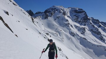 A skier wanders through a snowy mountain landscape. In the background, majestic mountains can be seen under a clear blue sky.